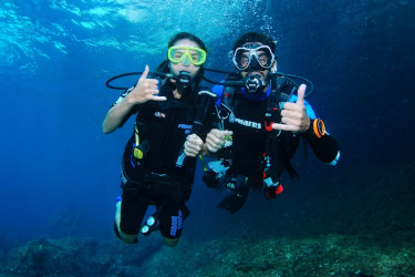 Two scuba divers underwater giving the “shaka” hand sign, smiling at the camera in clear blue water