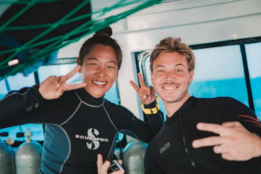 Two scuba divers smiling and making peace signs on a dive boat before entering the water