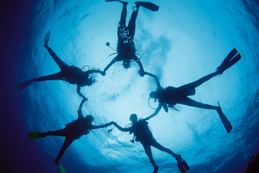 Silhouettes of five scuba divers holding hands in a circle viewed from below