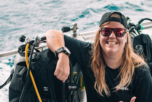 Smiling dive instructor named Sal sitting on a boat next to scuba gear, wearing sunglasses and a cap