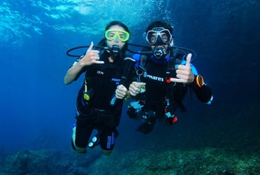 Two scuba divers underwater giving the “shaka” hand sign, smiling at the camera in clear blue water