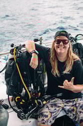 Smiling dive instructor named Sal sitting on a boat next to scuba gear, wearing sunglasses and a cap